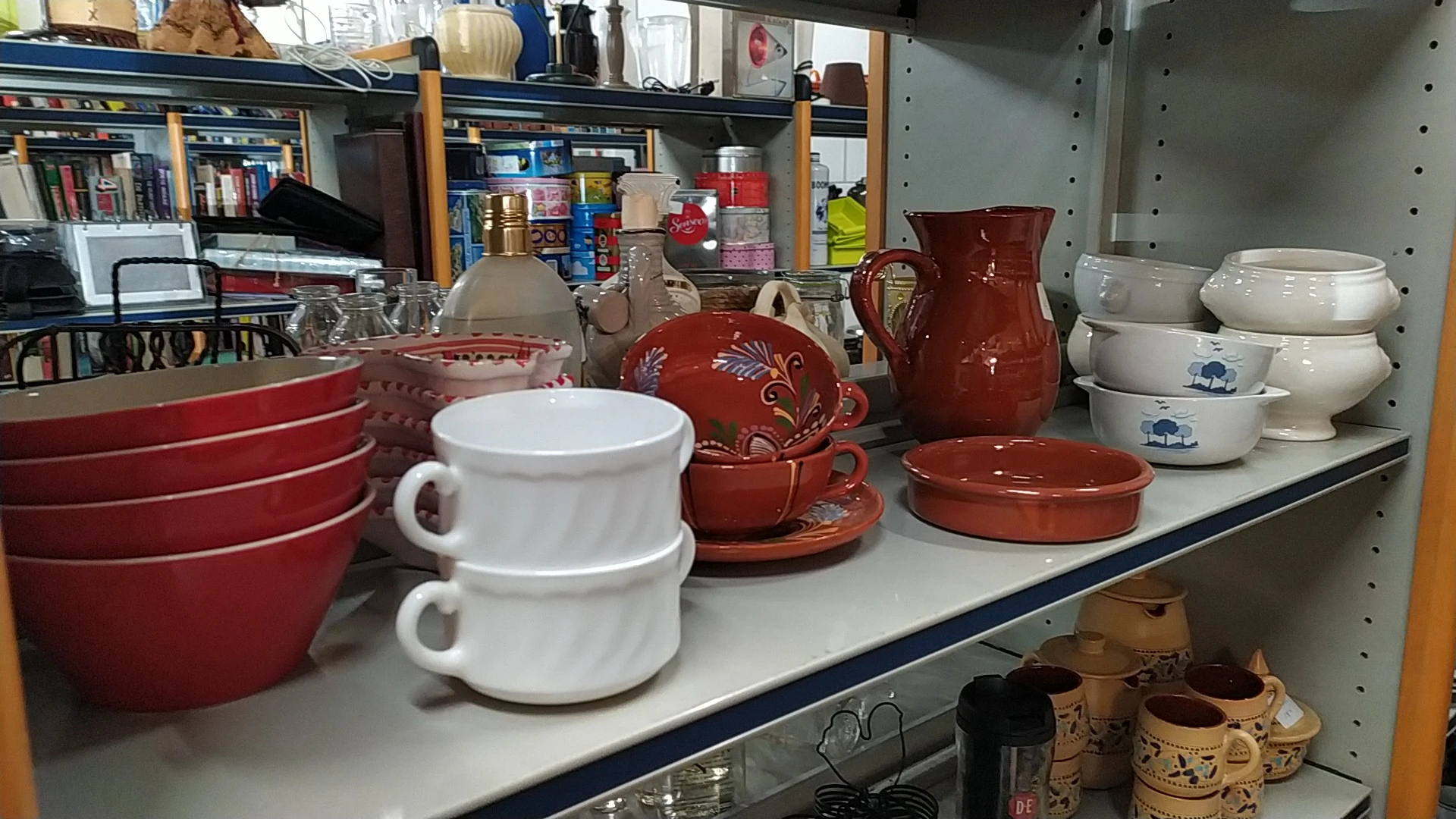 Uitgestalde serviesgoed en huishoudartikelen op een toonbank, waaronder witte kopjes, rode schalen en potten met decoraties