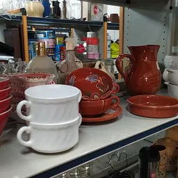 Uitgestalde serviesgoed en huishoudartikelen op een toonbank, waaronder witte kopjes, rode schalen en potten met decoraties