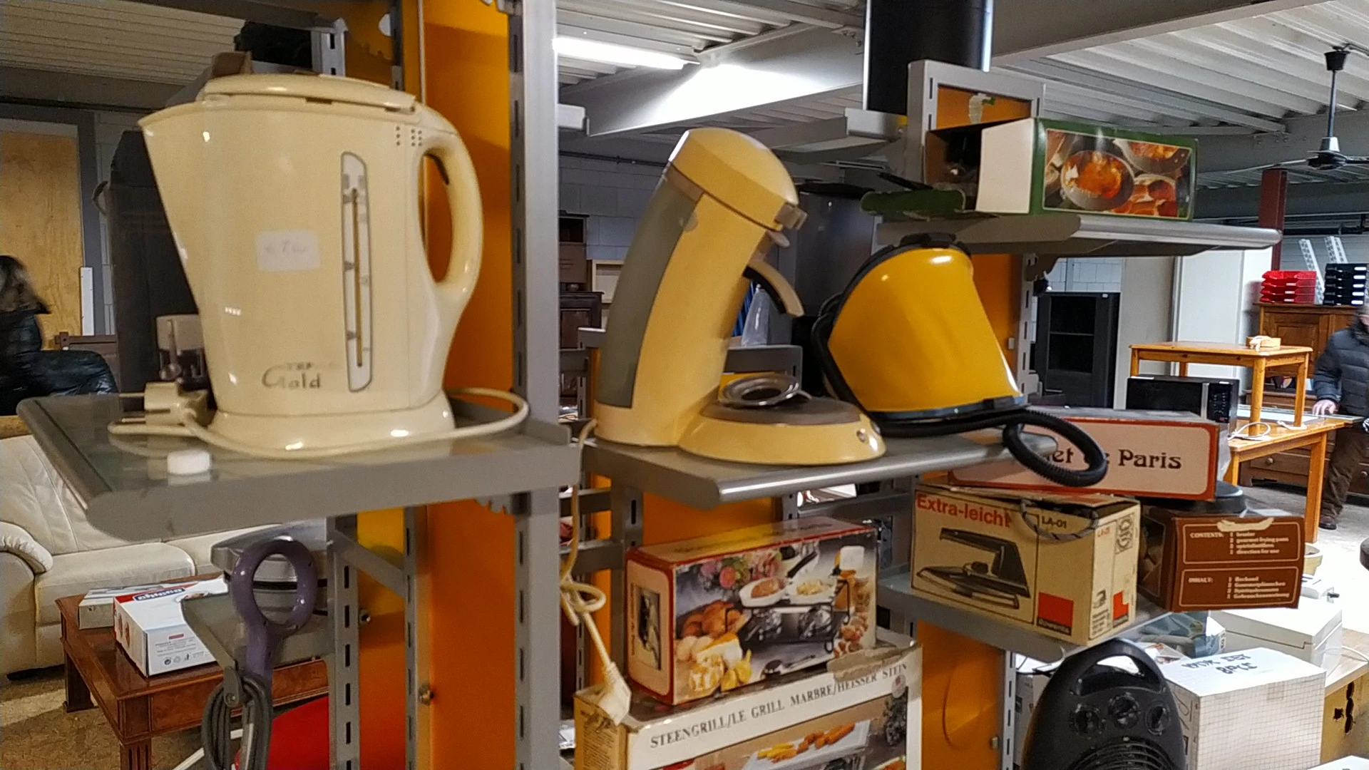 Verzameling tweedehands huishoudapparaten op schappen: waterkokers, koffiezetapparaten en andere keukengerei in geel, wit en 