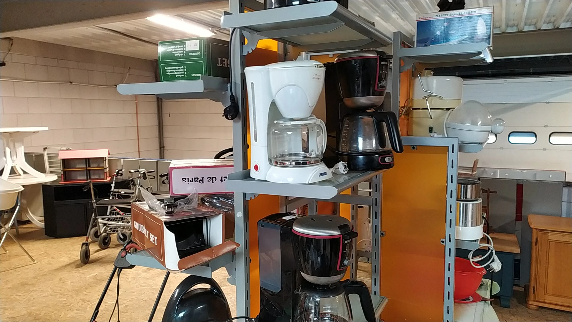 Keukenapparatuur op metalen stellingkasten, waaronder koffiezetapparaten en een rode ketel