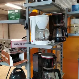 Keukenapparatuur op metalen stellingkasten, waaronder koffiezetapparaten en een rode ketel