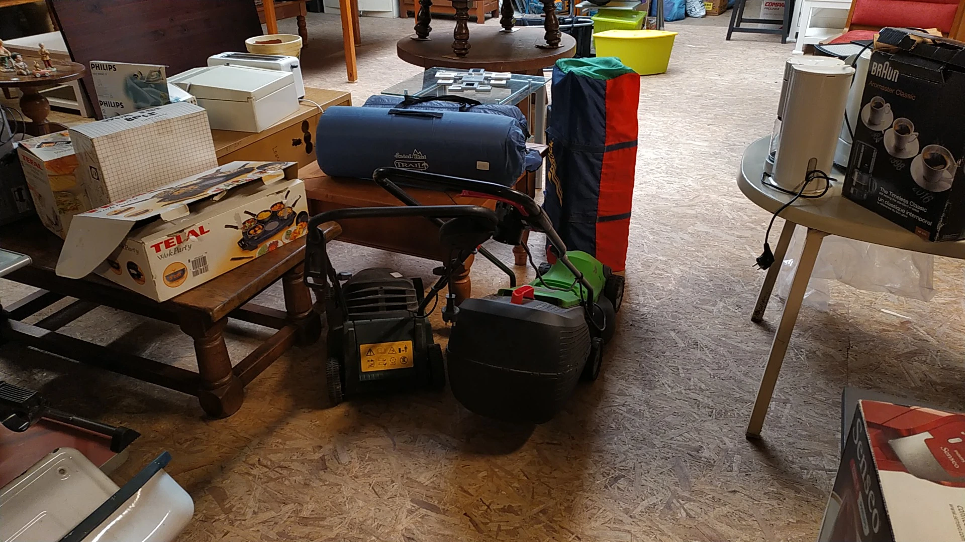 Winkelvloer met diverse tweedehands spullen en meubels, waaronder een grill, opbergkisten en huishoudartikelen.