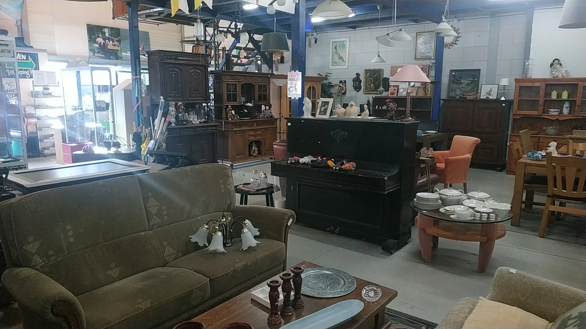 Winkelvloer met meubels waaronder bank, piano, fauteuil en salontafels, stellingen met diverse spullen op achtergrond