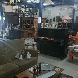 Winkelvloer met meubels waaronder bank, piano, fauteuil en salontafels, stellingen met diverse spullen op achtergrond