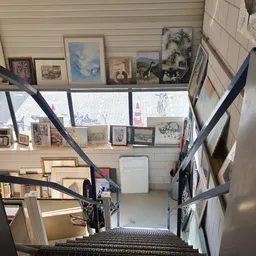 Overzicht van een kringloopwinkel met trappen, diverse kunstwerken en ingelijste afbeeldingen aan muren en stellingen