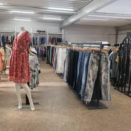 Binnenaanzicht van kringloopwinkel met rekken vol tweedehands kleding, pop met rode jurk en fluorescente plafondverlichting
