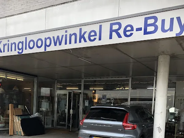 Gevel van Kringloopwinkel Re-Buy met glazen deuren, parkeerplaats en auto's ervoor