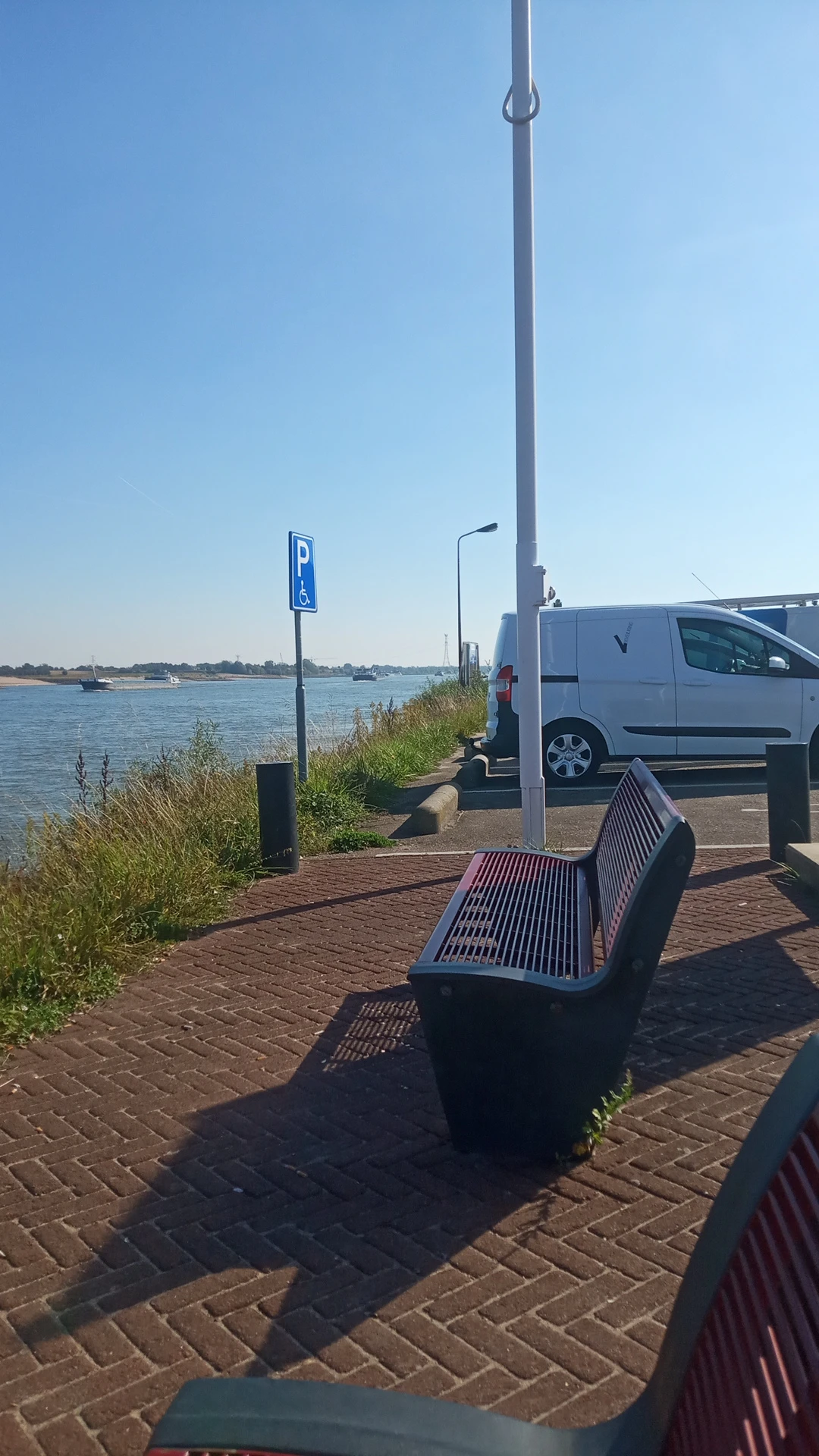 Parkeerplaats langs water met witte bestelwagen, parkeerbordje en houten bankje