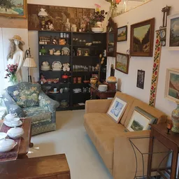 Kringloopwinkelinterieur met beige bank, zwarte vitrinekast vol serviesgoed en decoraties, schilderijen en klok aan de muur