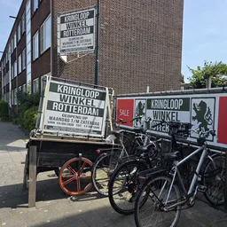 Voorgevel van kringloopwinkel met etalage, fietsen geparkeerd ervoor en reclameborden