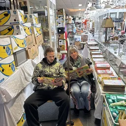 Twee personen zitten in een kringloopwinkel en bladeren door boeken, omringd door rekken met boeken en speelgoed