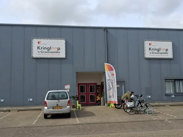 Grijze gevel van kringloopwinkel met twee borden met naam Kringloop 's-Gravenpolder, rode deuren, witte auto en fietsen gepar