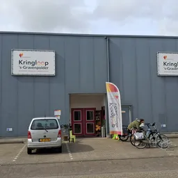 Grijze gevel van kringloopwinkel met twee borden met naam Kringloop 's-Gravenpolder, rode deuren, witte auto en fietsen gepar