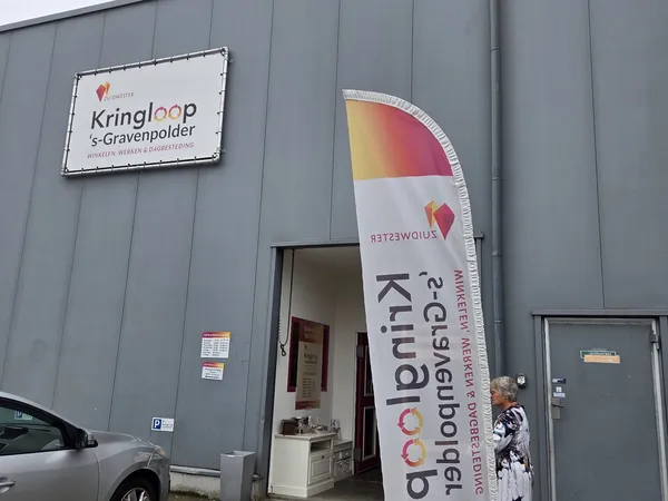 Grijze gevel van kringloopwinkel met witte naambordje 'Kringloop s-Gravenpolder' en opgeklapte windzak met logo en website