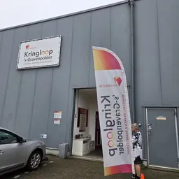 Grijze gevel van kringloopwinkel met witte naambordje 'Kringloop s-Gravenpolder' en opgeklapte windzak met logo en website