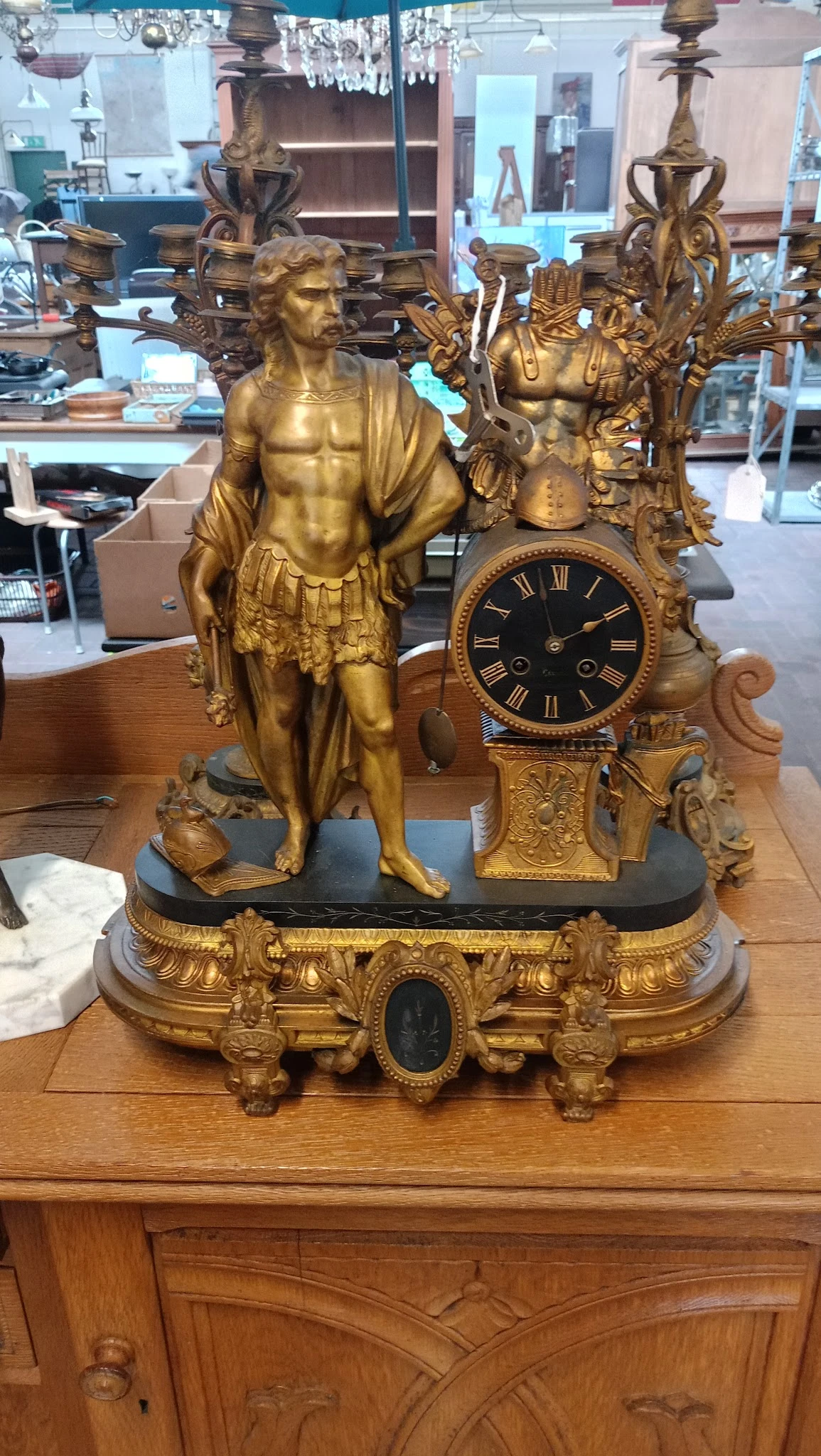 Gouden decoratief mantelklok met beeldhouwwerk van een figuur in klassieke stijl op zwart marmer voetstuk