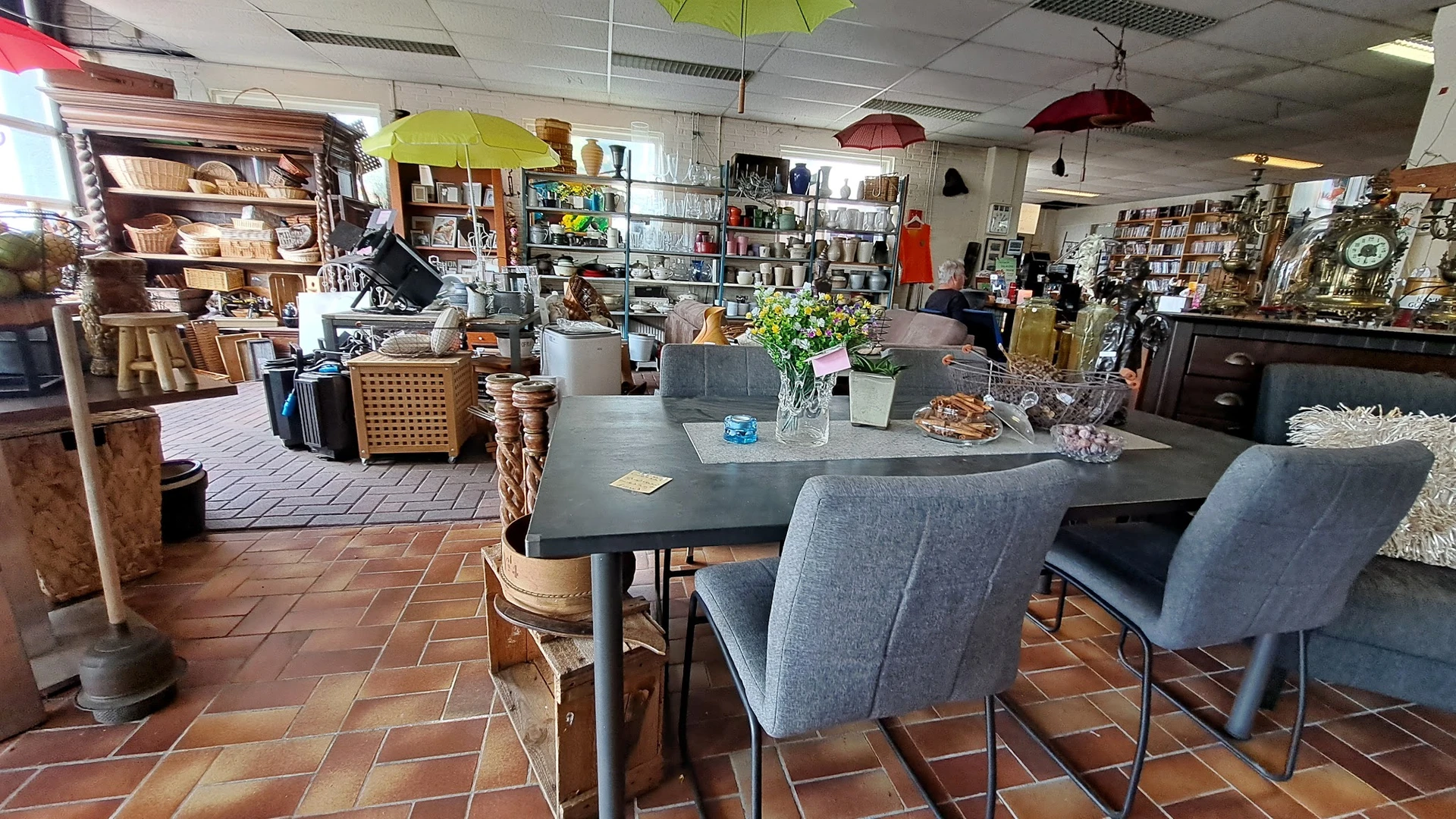 Winkelvloer met zwarte eettafel, grijze stoelen, gele en rode hangende paraplus, stellingen en diverse inrichtingsartikelen