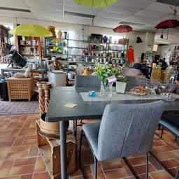 Winkelvloer met zwarte eettafel, grijze stoelen, gele en rode hangende paraplus, stellingen en diverse inrichtingsartikelen