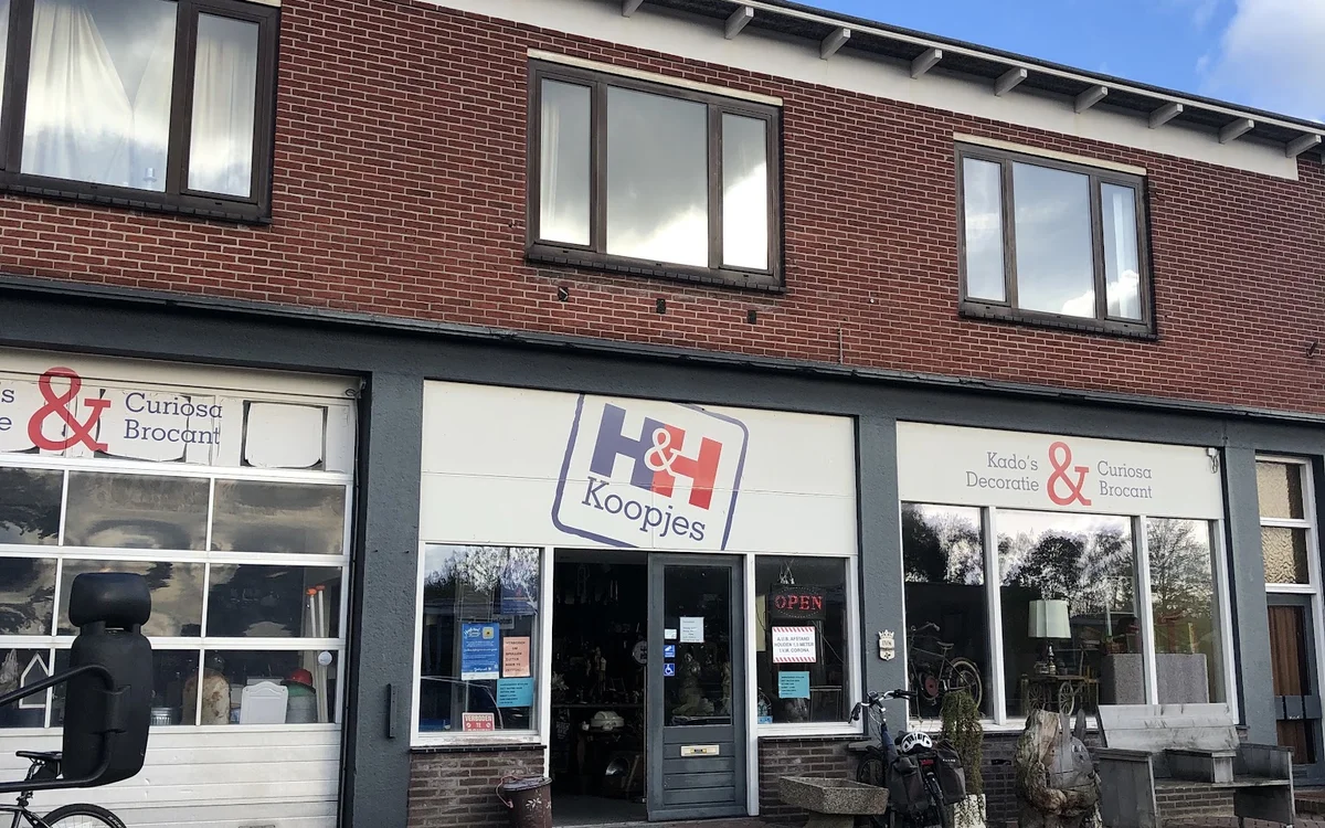Voorgevel van kringloopwinkel met bruine bakstenen, grote ramen en donkerblauwe deuren, fietsen ervoor