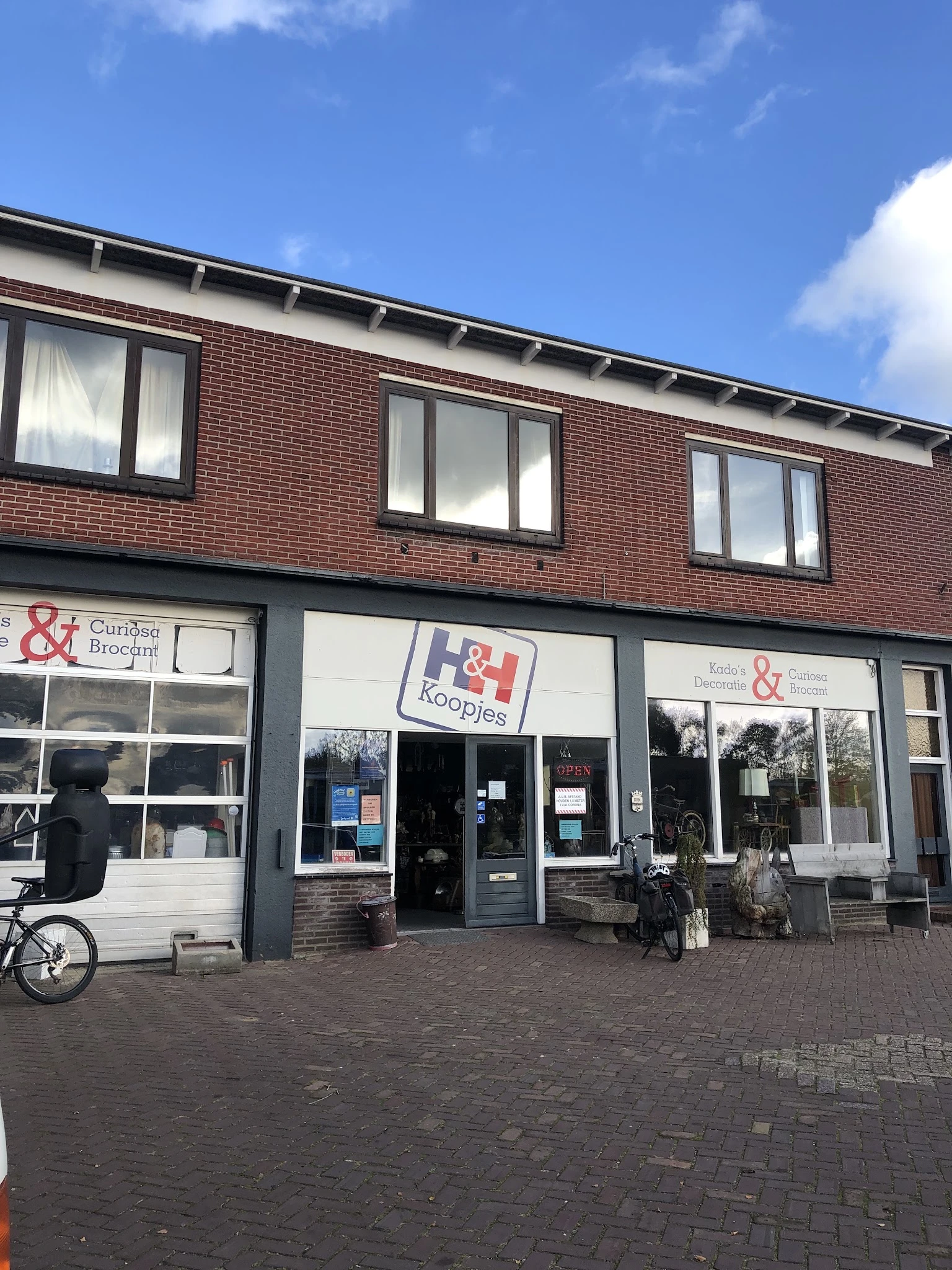 Voorgevel van kringloopwinkel met bruine bakstenen, grote ramen en donkerblauwe deuren, fietsen ervoor