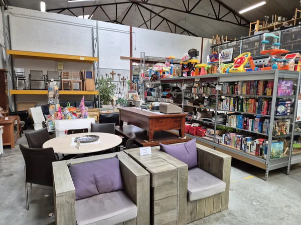 Winkelvloer met zithoeken met paarse kussens, ronde tafel, en rekken vol boeken en spullen tegen de achterste wand