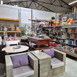 Winkelvloer met zithoeken met paarse kussens, ronde tafel, en rekken vol boeken en spullen tegen de achterste wand