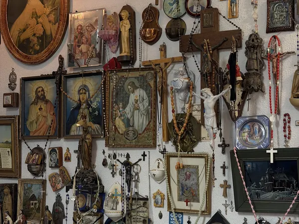 Verzameling religieuze voorwerpen en kunstvoorwerpen aan een witte muur, waaronder ikonen, kruisen, beelden en devotionalia.