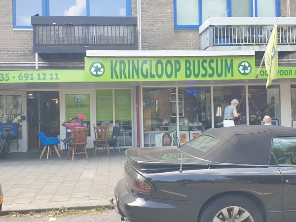 Gevel van kringloopwinkel met groene luifel en winkelnaam, terras ervoor met stoelen