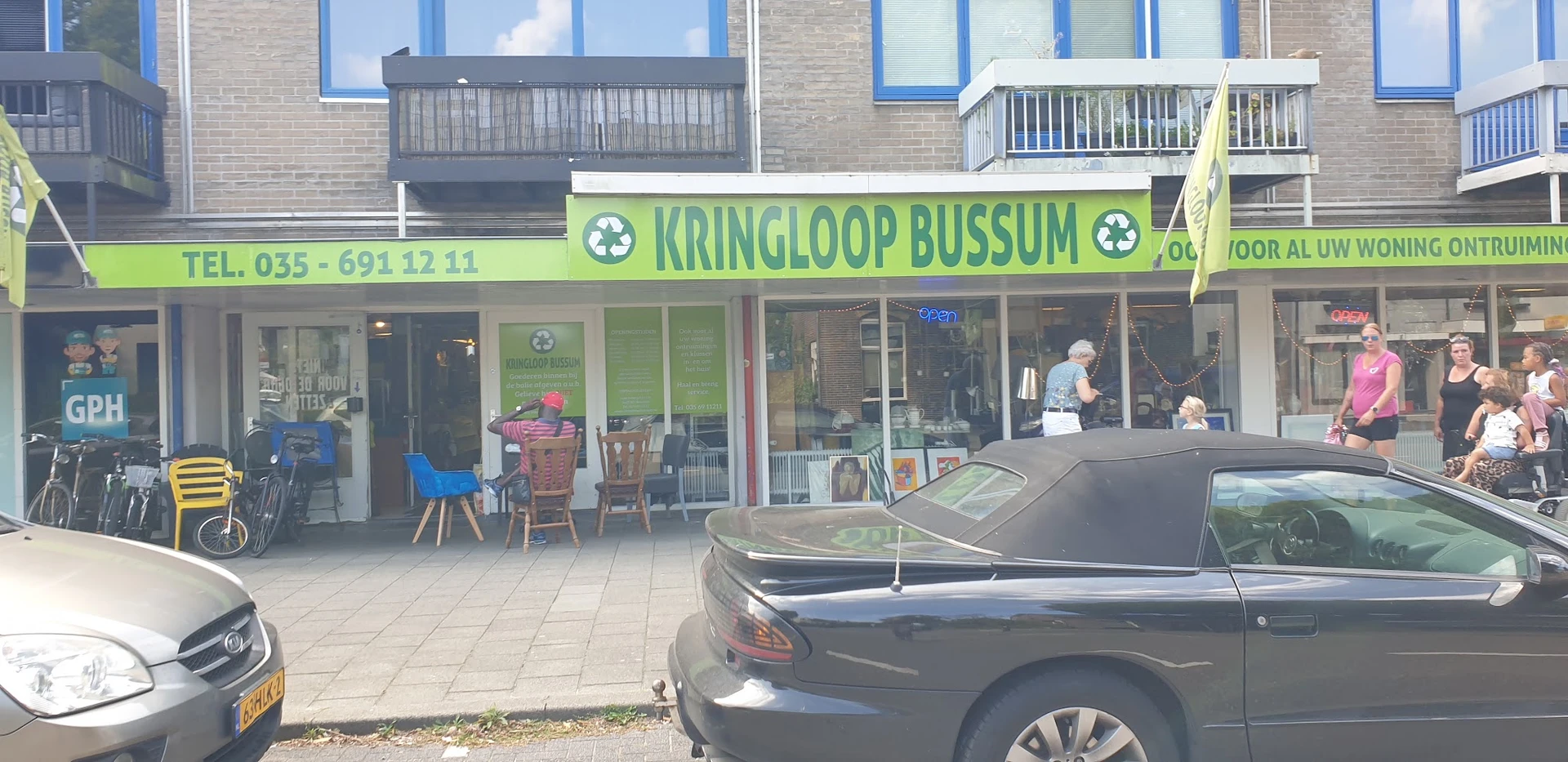 Gevel van kringloopwinkel met groene luifel en winkelnaam, terras ervoor met stoelen