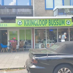 Gevel van kringloopwinkel met groene luifel en winkelnaam, terras ervoor met stoelen