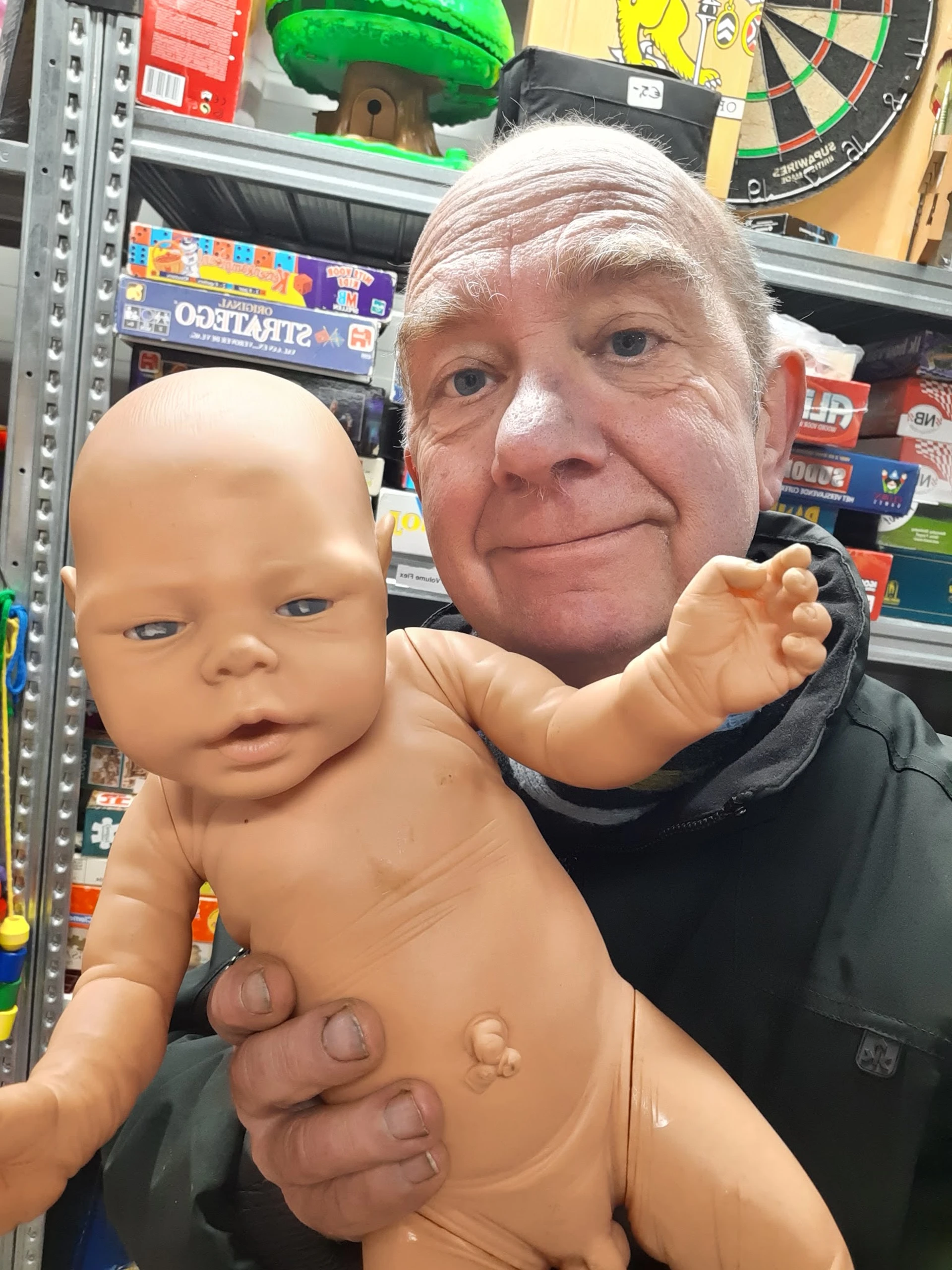 Een man houdt een naakte babypop vast in een kringloopwinkel