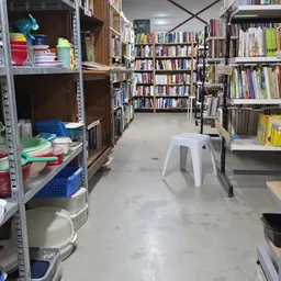 Binnenkant van kringloopwinkel met boekenrekken, stellingen met serviesgoed en huishoudartikelen, en witte plastic stoel op b