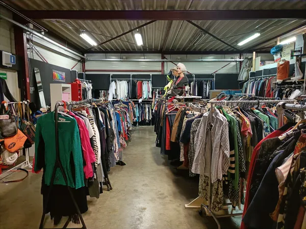 Overzicht van de kringloopwinkelvloer met meerdere rekken vol tweedehands kleding, verlicht door fluorescente lampen.