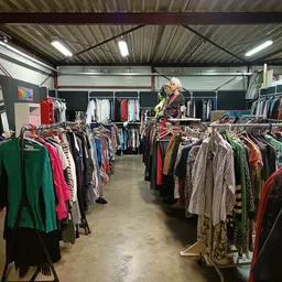 Overzicht van de kringloopwinkelvloer met meerdere rekken vol tweedehands kleding, verlicht door fluorescente lampen.