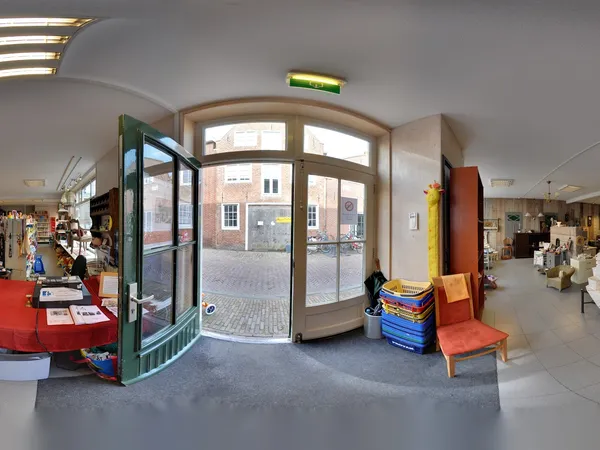 Entreehal van kringloopwinkel met glazen deuren, rode tafel met spullen links, en uitzicht op straat