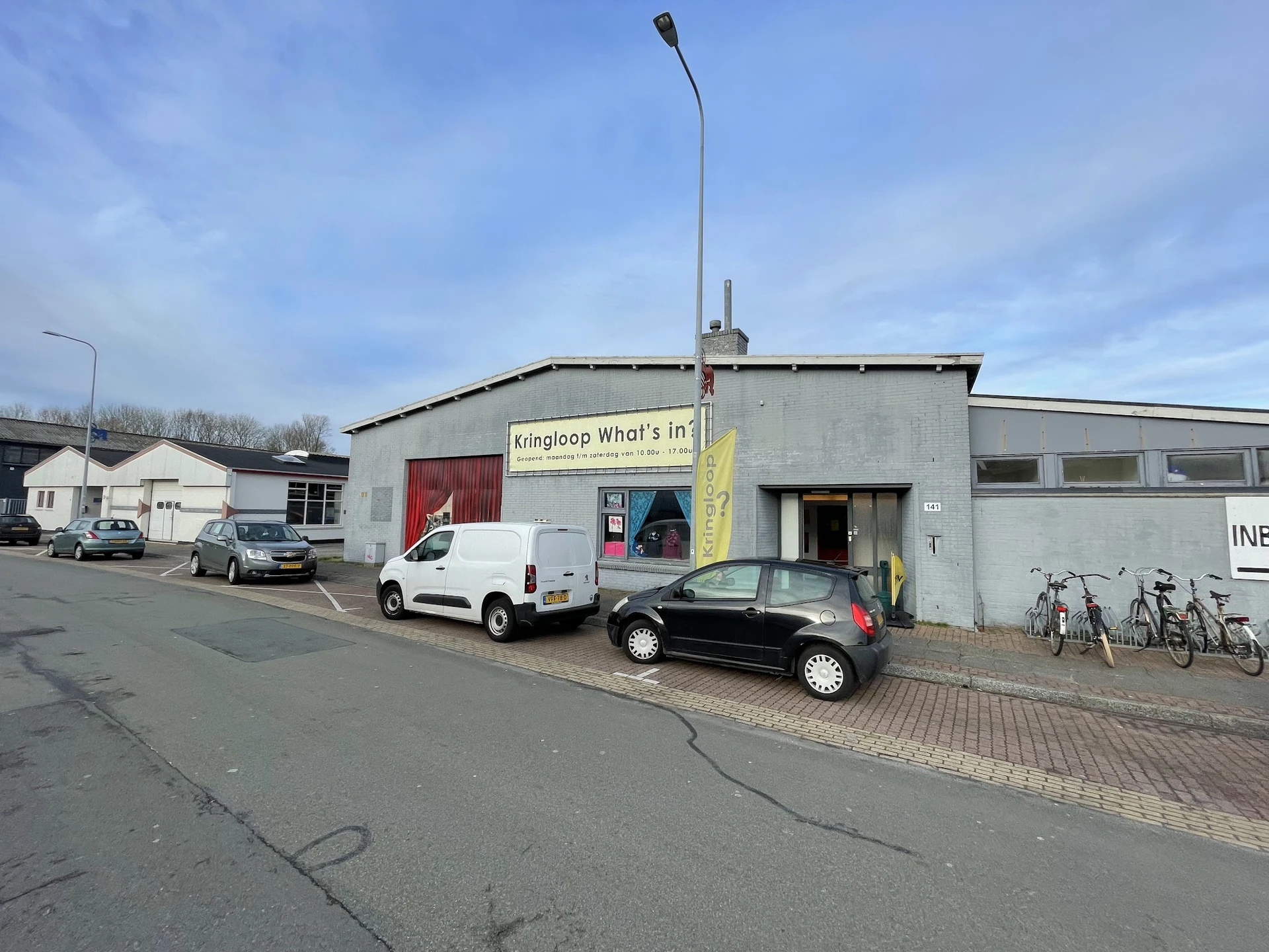 Gevel van kringloopwinkel What's in? met gele banner, geparkeerde auto's en fietsen ervoor