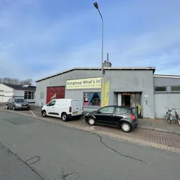 Gevel van kringloopwinkel What's in? met gele banner, geparkeerde auto's en fietsen ervoor