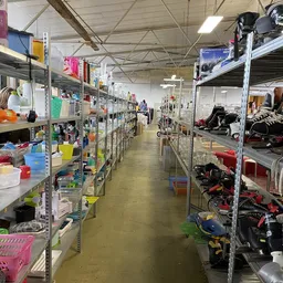 Lange gang met metalen stellingen vol huishoudelijke spullen, speelgoed en diverse gebruiksartikelen aan beide zijden