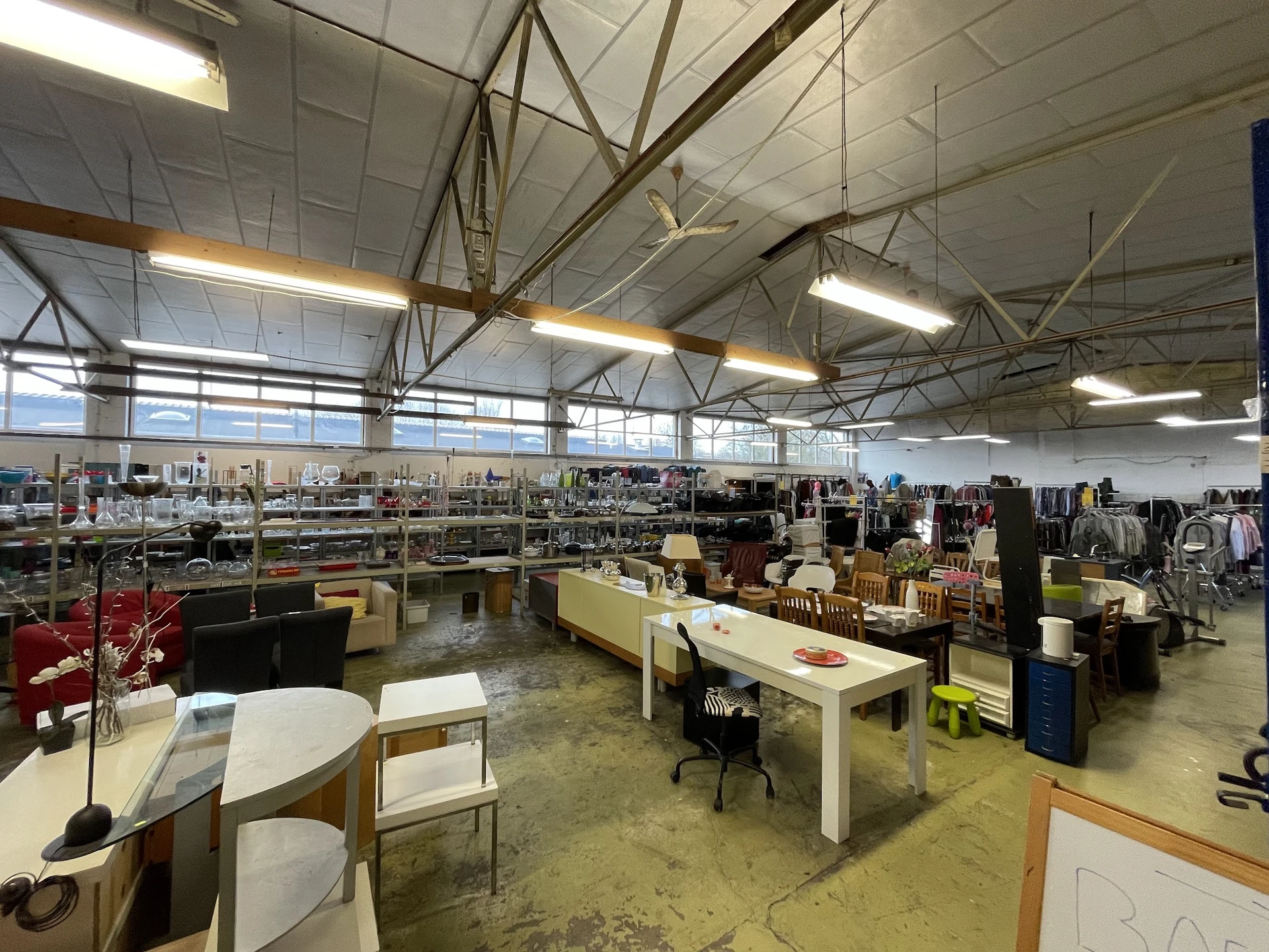 Ruim magazijn- of winkelvloer met verschillende afdelingen, tafels met spullen, rekken met kleding en diverse tweedehandsarti