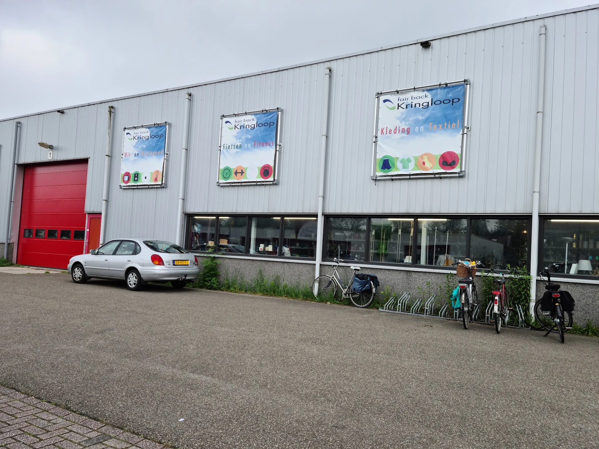 Wit golfplaten pand met rode deur, winkelramen en uitgestalde fietsen op het parkeerterrein