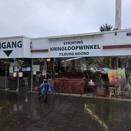 Gevel van kringloopwinkel met ingang, groot raam en houten deur, naam duidelijk zichtbaar, fietser op straat