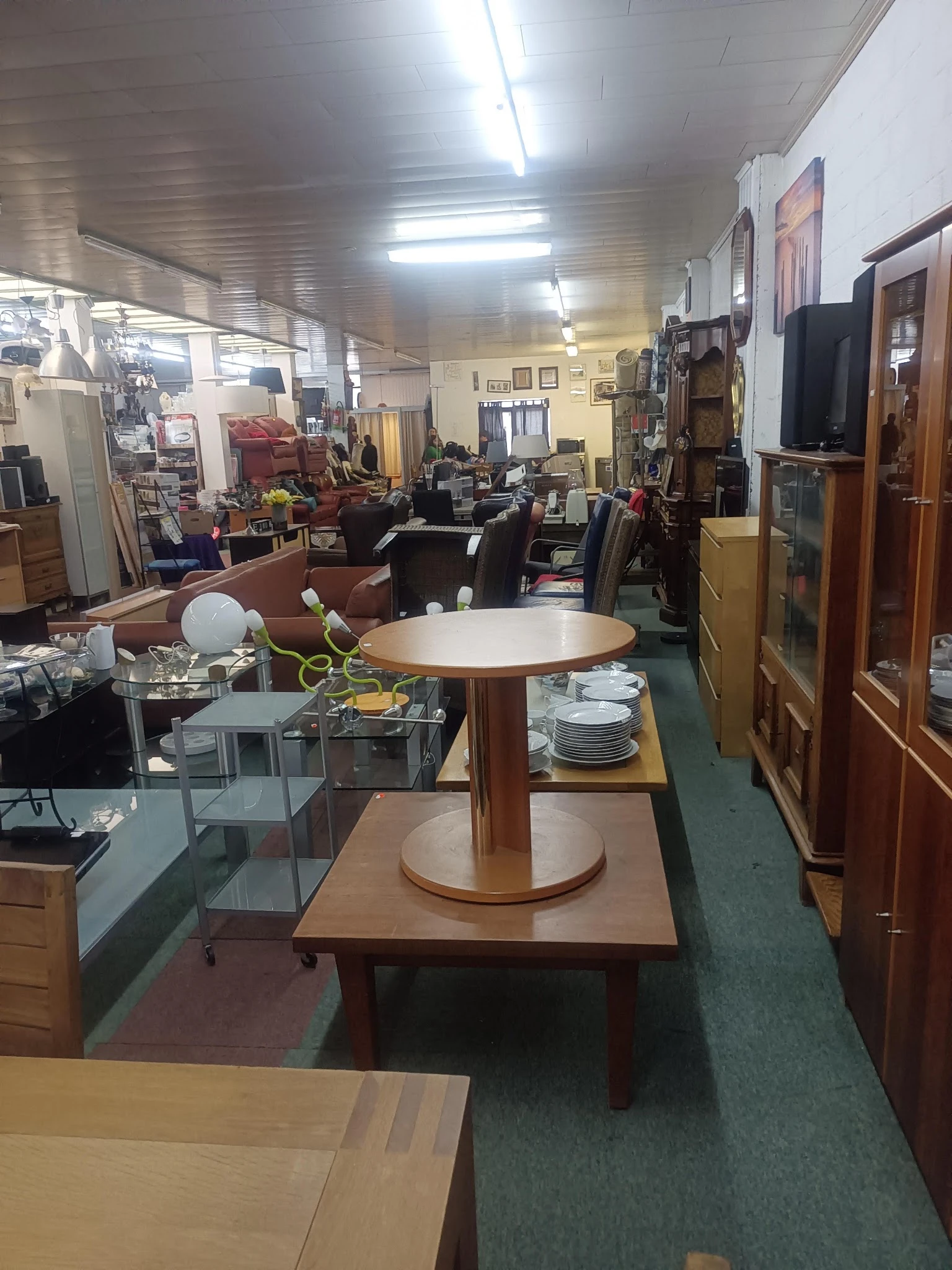 Winkelvloer van kringloopwinkel met ronde tafel, serviesgoed, meubels en verschillende afdelingen op achtergrond