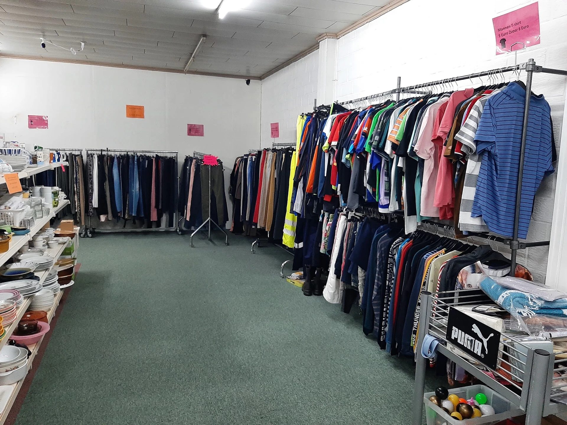 Kringloopwinkel met kledingrekken, stellingen en diverse tweedehands artikelen op groene vloer