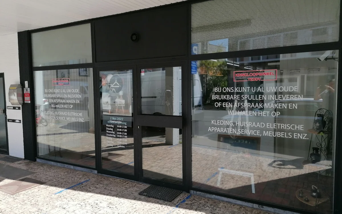 Glazen pui van kringloopwinkel met zwarte frame en witte tekst op ramen, ingang zichtbaar