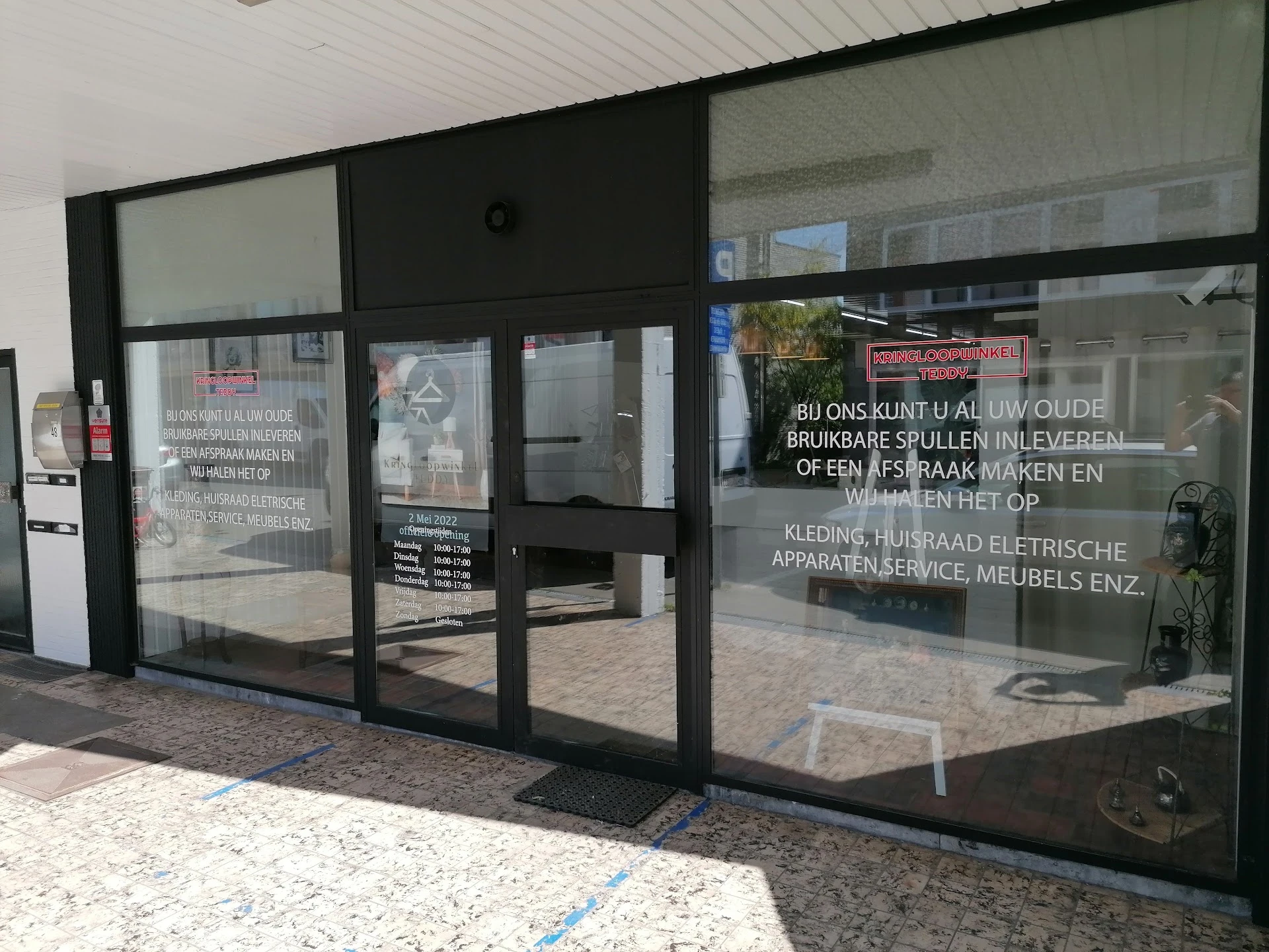 Glazen pui van kringloopwinkel met zwarte frame en witte tekst op ramen, ingang zichtbaar