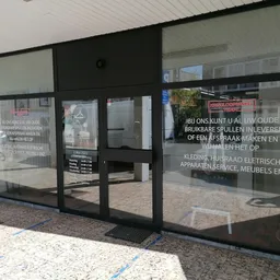 Glazen pui van kringloopwinkel met zwarte frame en witte tekst op ramen, ingang zichtbaar