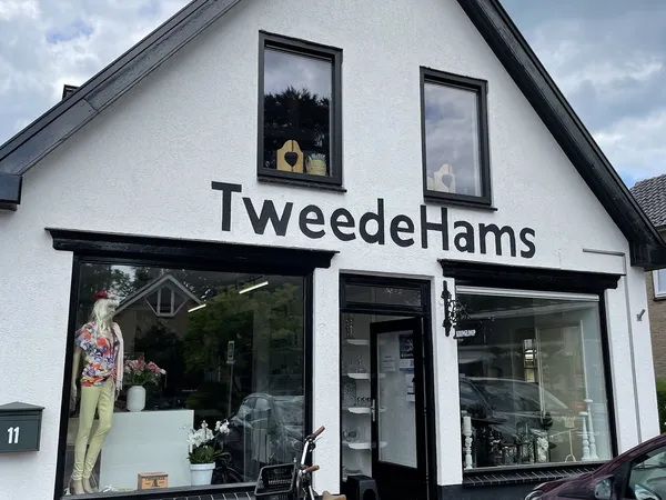 Witte gevel van kringloopwinkel met zwarte ramen en naam TweedeHams, mannequin in etalage