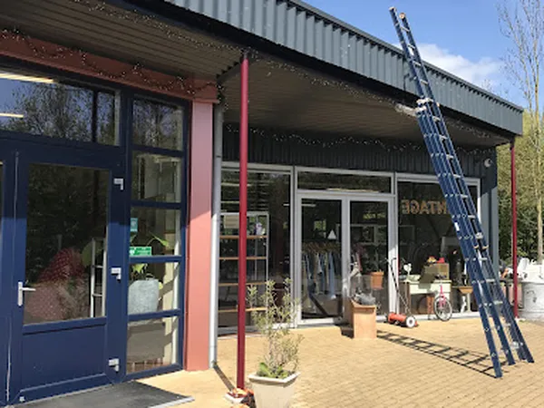 Voorkant van kringloopwinkel met blauwe deur, rode zuilen en glazen puien, ladder tegen gevel