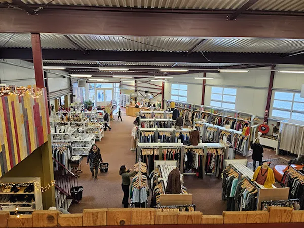 Ruim magazijn met kledingrekken, tafels met artikelen en winkelende klanten op houten vloer