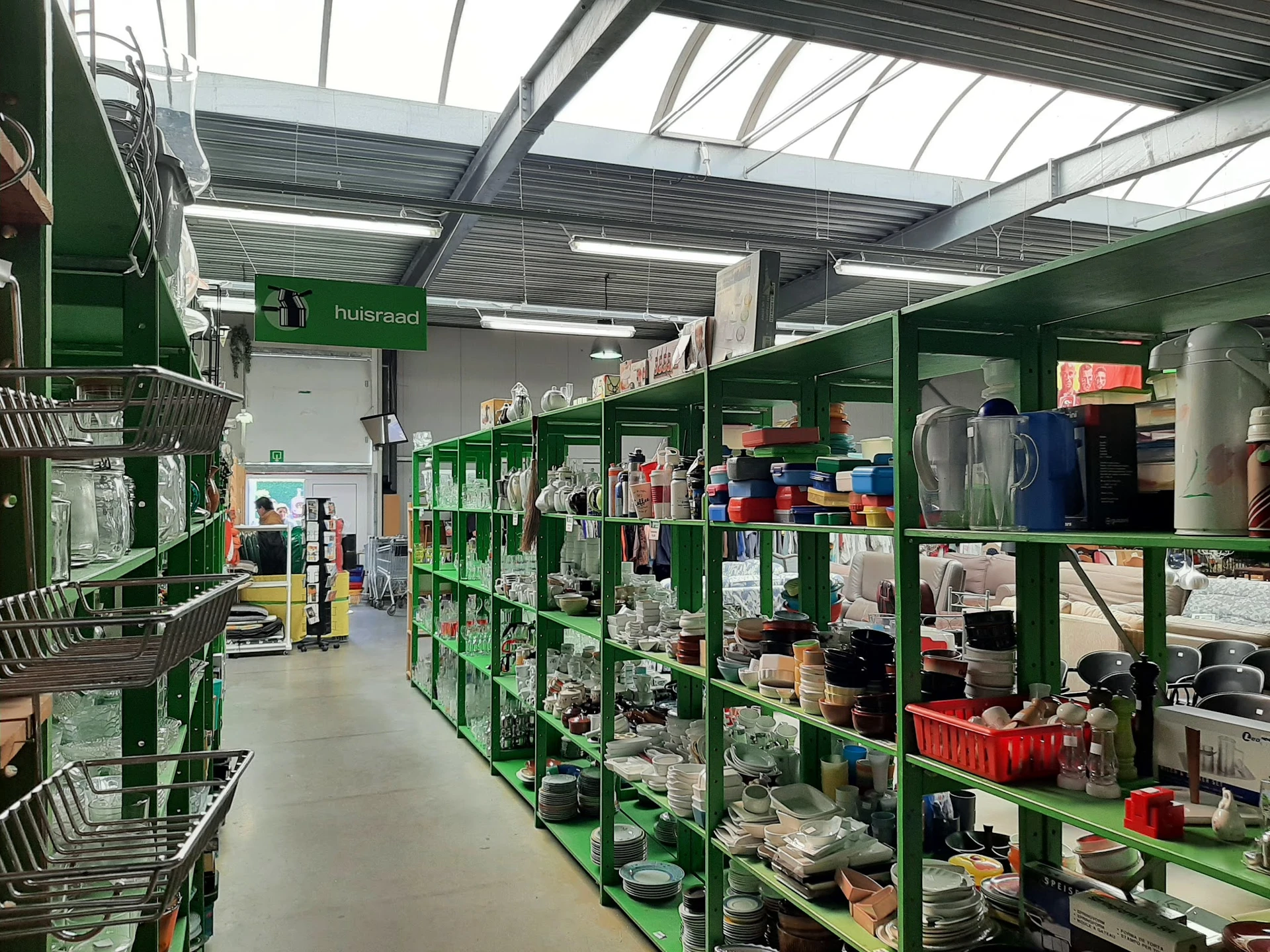 Winkelvloer met groene stellingen vol tweedehands huishoudartikelen, kleding en diverse goederen in een kringloopwinkel